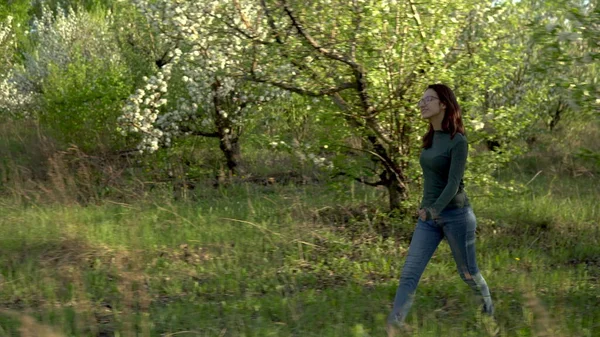 Młoda kobieta przechodzi przez kwitnący sad jabłkowy. Widok przez drzewa. — Zdjęcie stockowe