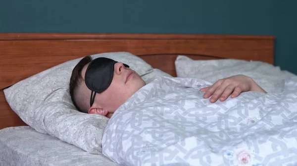 A young man sleeps in a sleep mask. A man lies in bed in his room.