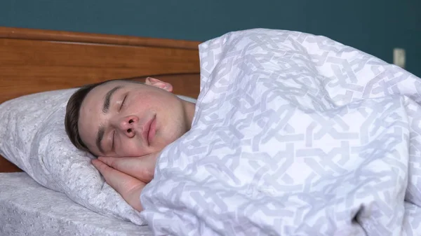 Um jovem está a dormir debaixo dos lençóis. Um homem jaz na cama em seu quarto . — Fotografia de Stock