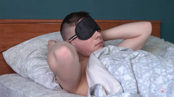 O jovem vai para a cama e coloca uma máscara. Um homem jaz na cama em seu quarto . — Fotografia de Stock