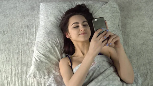 La joven se despertó y cogió el teléfono. Una mujer sacó un smartphone de debajo de las almohadas. Vista desde arriba . —  Fotos de Stock