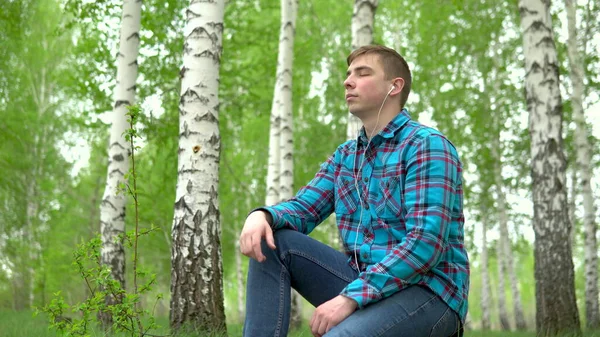 Молодий чоловік сидить у природі з навушниками у вухах. Чоловік сидить на пні в березовому лісі і слухає музику . — стокове фото