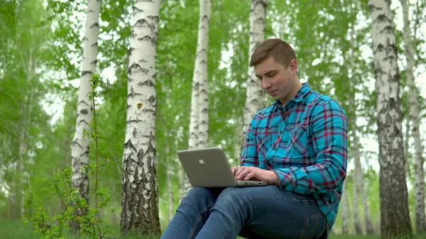Jovem na natureza com um laptop nas mãos. Um homem senta-se em um toco em uma floresta de vidoeiro e leva obras através de um netbook . — Fotografia de Stock