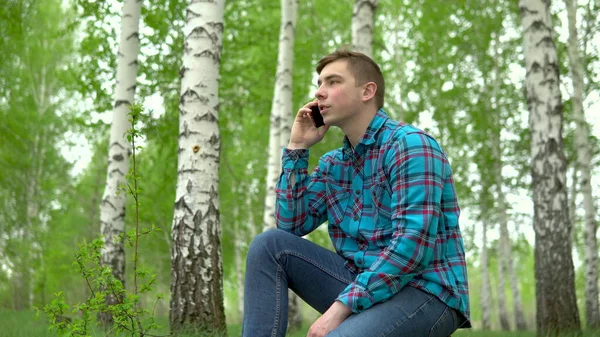 Молодой человек в природе с телефоном. Мужчина сидит на пне в березовом лесу и разговаривает по телефону . — стоковое фото