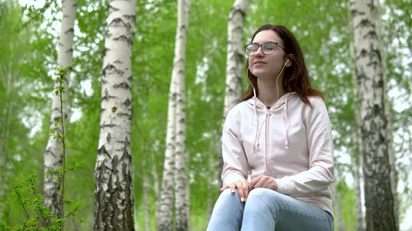 Mladá žena sedí v přírodě se sluchátky v uších. Dívka sedí na pařezu v břízovém lese a poslouchá hudbu. — Stock fotografie