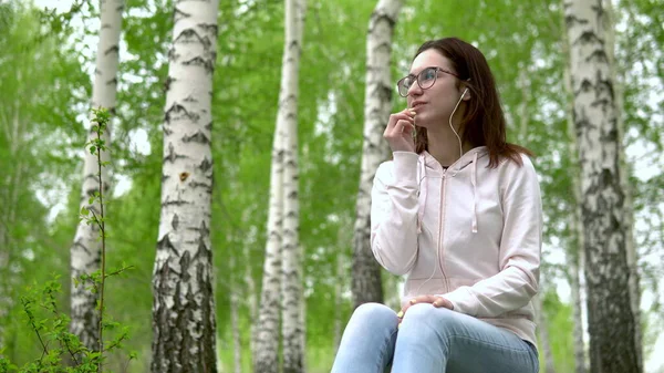Egy fiatal nő a természetben fejhallgatón keresztül beszél telefonon. Egy lány ül egy fatönkön egy nyírfa erdőben fejhallgatóval. — Stock Fotó