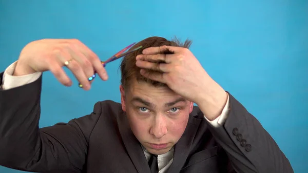Podnikatel si stříhá vlasy na modrém pozadí. Muž v obleku se zastřihl nůžkami.. — Stock fotografie