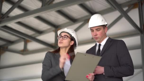 De man keurt het project goed. Jonge man en vrouw in helmen met documenten op een bouwplaats. De bazen in pakken bespreken een architectuurproject. Mensen kijken om zich heen. — Stockvideo