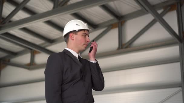 Un jeune homme portant un casque parle sur un talkie-walkie sur un chantier de construction. Le patron en costume regarde autour de lui.. — Video