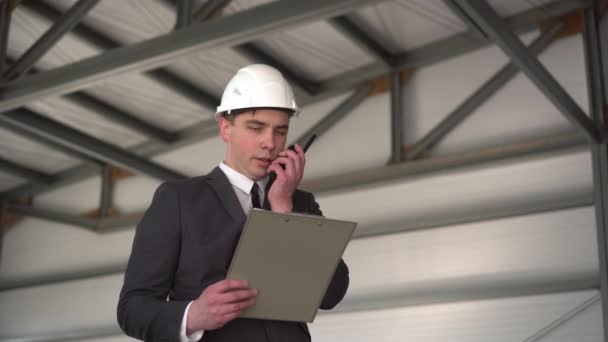 Un jeune homme portant un casque avec des documents parle sur un talkie-walkie sur un chantier de construction. Le patron en costume garde des documents sur l'architecture. Un homme regarde autour de lui. — Video