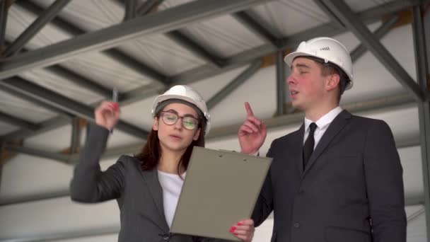 Jonge man en vrouw in helmen met documenten op een bouwplaats. De bazen in pakken bespreken een architectuurproject. Een man spreekt op een walkietalkie. Mensen kijken om zich heen. — Stockvideo