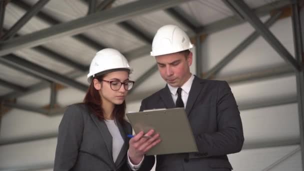 Jonge man en vrouw in helmen met documenten op een bouwplaats. De bazen in pakken bespreken een architectuurproject. De man is nerveus. Mensen kijken om zich heen. — Stockvideo