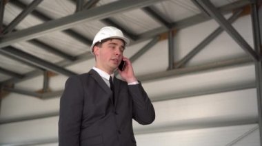 Kasklı genç bir adam inşaat alanında telefonla konuşuyor. Takım elbiseli patron telefonda konuşuyor..