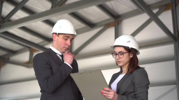 Jonge man en vrouw in helmen met documenten op een bouwplaats. Chiefs in pakken bespreken het architectuurproject. Mensen kijken om zich heen. — Stockvideo