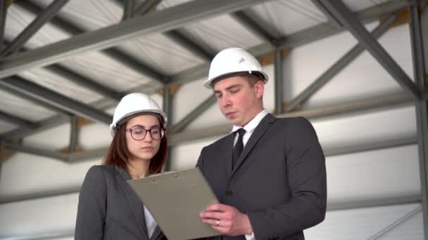 Joven hombre y mujer en cascos con documentos en un sitio de construcción. Jefes en trajes discutiendo el proyecto de arquitectura. — Vídeos de Stock