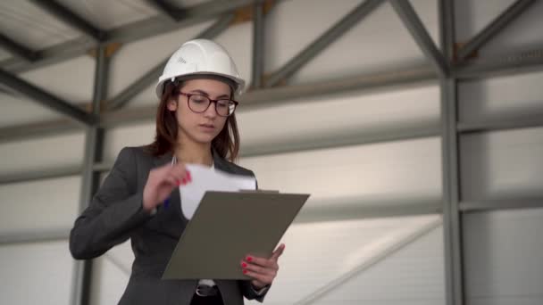 Jonge vrouw in een helm met documenten op een bouwplaats. De baas vrouw in een pak houdt documenten bij over architectuur. Het meisje kijkt om zich heen. — Stockvideo