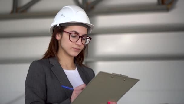 Jonge vrouw in een helm met documenten op een bouwplaats. De baas vrouw in een pak houdt documenten bij over architectuur. Het meisje kijkt om zich heen. — Stockvideo