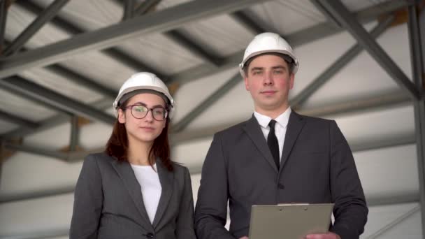 Jonge man en vrouw in helmen op een bouwplaats. Zakenmensen in pakken kijken naar de camera en tonen een duim.. — Stockvideo