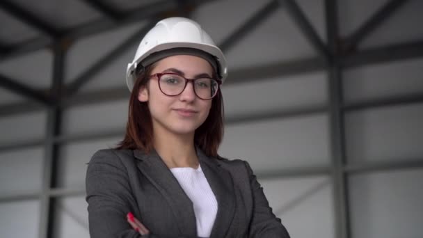 Een jonge vrouw met een beschermhelm kruiste haar armen en staat op een bouwplaats. De baas vrouw in een pak kijkt naar de camera. — Stockvideo