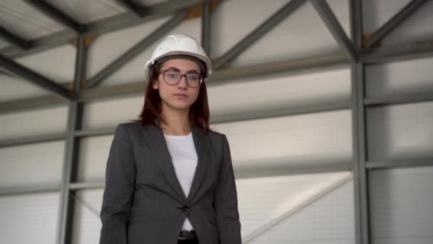 Una joven con un casco protector muestra un pulgar en una obra de construcción. La jefa con traje mira a la cámara. — Vídeo de stock