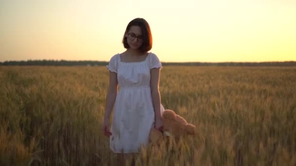 Młoda kobieta idzie przez pole pszenicy z misiaczkiem o zachodzie słońca. Dziewczyna trzyma misia w ręku widok z przodu. — Wideo stockowe