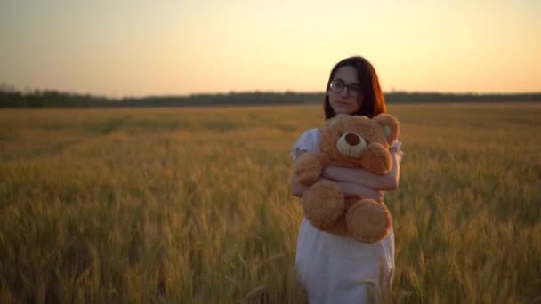 En ung kvinna går genom ett vetefält med en nalle vid solnedgången. Flicka kramar en teddybjörn i händerna framifrån. — Stockvideo