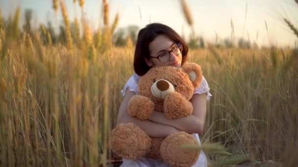 A young woman sits in a dried wheat field with a teddy bear. Girl hugs a teddy bear in hands front view. — Stock Video