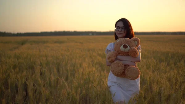 Une jeune femme marche dans un champ de blé avec un ours en peluche au coucher du soleil. Fille étreint un ours en peluche dans les mains vue de face. — Photo