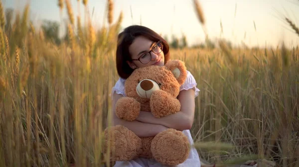 Mladá žena sedí na vysušeném pšeničném poli s medvídkem. Dívka objetí a medvídek v ruce přední pohled. — Stock fotografie
