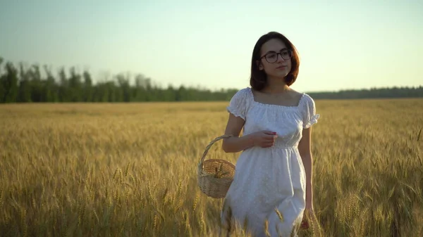 A young woman is walking on a yellow wheat field with a basket in her hands. Straw basket with spikelets of wheat. Front view. — Stock Photo, Image