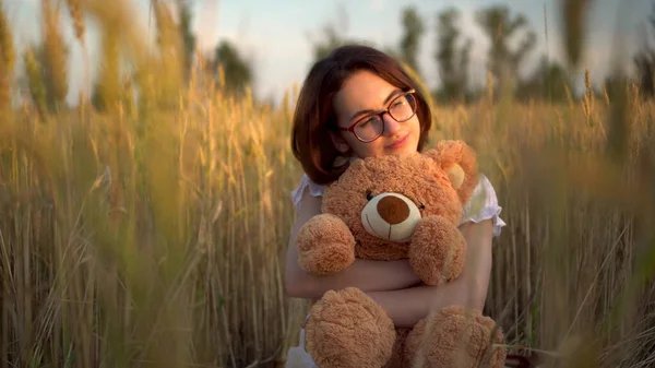 Eine junge Frau sitzt mit einem Teddybär in einem getrockneten Weizenfeld. Mädchen umarmt einen Teddybär in den Händen. — Stockfoto