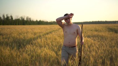 Gömleksiz genç bir adam elinde bir sopayla buğday tarlası boyunca yürüyor. Gün batımında sarı tarlada bir adam.