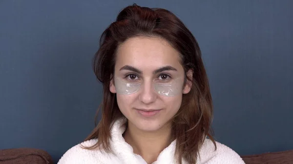 Wanita muda menempatkan tambalan di wajahnya. Biru hydrogel patch untuk meremajakan kulit wajah. Gadis yang duduk di mantel putih. Tutup. — Stok Foto