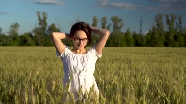Młoda kobieta w białej sukience stoi na zielonym polu pszenicy i dotyka jej włosów. Dziewczyna cieszy się słońcem w terenie. — Wideo stockowe