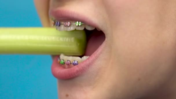 Девочка-подросток с брекетами на зубах ест сельдерей на синем фоне. Девушка с цветными брекетами откусывает стебель сельдерея крупным планом. — стоковое видео