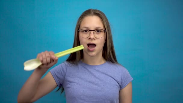 Ragazza adolescente con l'apparecchio sui denti mangia sedano su uno sfondo blu. Una ragazza con l'apparecchio colorato morde un gambo di sedano. — Video Stock