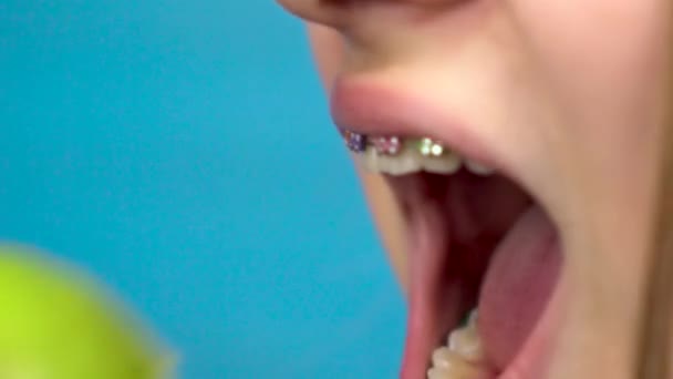 Teenager girl with braces on her teeth eats a green apple on a blue background. Girl with colored braces bites off an apple closeup. — Stock Video