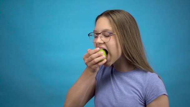 Tini lány fogszabályzóval a fogán eszik egy zöld almát, kék háttérrel. A színes fogszabályzós lány leharap egy almát.. — Stock videók