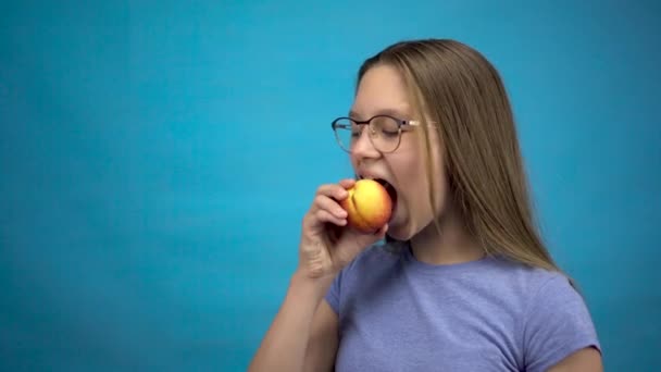 Chica adolescente con aparatos ortopédicos en los dientes se come un melocotón sobre un fondo azul. Chica con frenos de colores muerde de un melocotón. — Vídeos de Stock