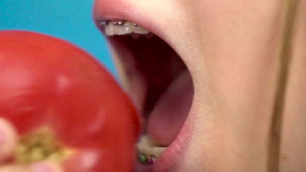 Teenager-Mädchen mit Zahnspange isst eine rote Tomate auf blauem Hintergrund. Mädchen mit farbigen Hosenträgern beißt Tomaten-Nahaufnahme ab. — Stockvideo