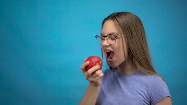 Dişlerinde diş teli olan genç bir kız mavi arka planda kırmızı domates yiyor. Renkli diş teli olan bir kız domatesi ısırıp koparır.. — Stok video