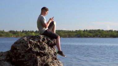 Genç bir adam elinde telefonla nehrin kıyısındaki bir taşın üzerinde oturuyor. Adam akıllı telefonuyla yakın çekimde mesaj atıyor..