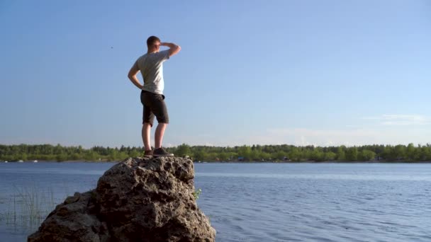 Een jongeman staat op een steen bij de rivier en bewondert het landschap. Een man in de natuur. — Stockvideo