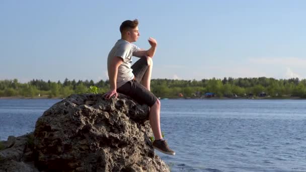 En ung man sitter på en sten vid floden och beundrar naturen. En man i naturen närbild. — Stockvideo