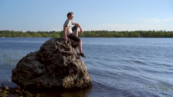 Un joven se sienta en una piedra junto al río y admira la naturaleza. Un hombre en la naturaleza. — Vídeo de stock