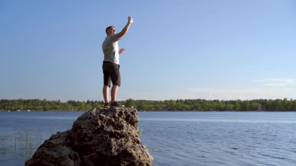 En ung man står på en sten och tar bilder på sig själv i telefonen. En man tar en selfie i naturen. — Stockvideo