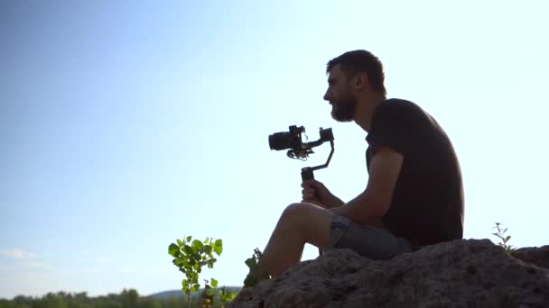 Mladý muž sedí na kameni s fotoaparátem v ruce. Muž střílí na přírodu kamery. — Stock video