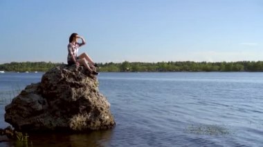 Genç bir kadın nehir kenarındaki bir taşın üzerinde oturur ve doğaya hayran kalır. Doğadaki kız.