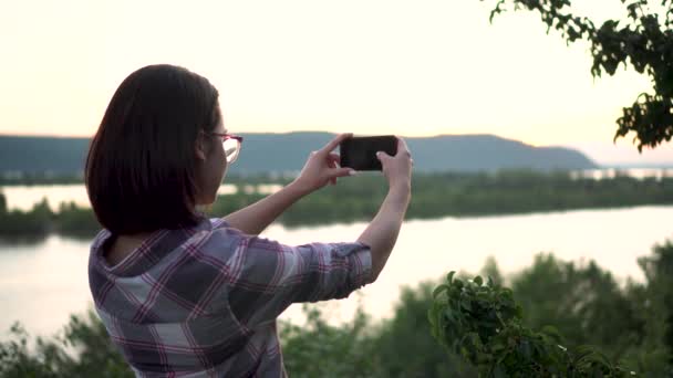 Uma jovem mulher está em uma colina contra o fundo do rio e montanhas e fotografa a paisagem. A menina tira fotos na natureza . — Vídeo de Stock