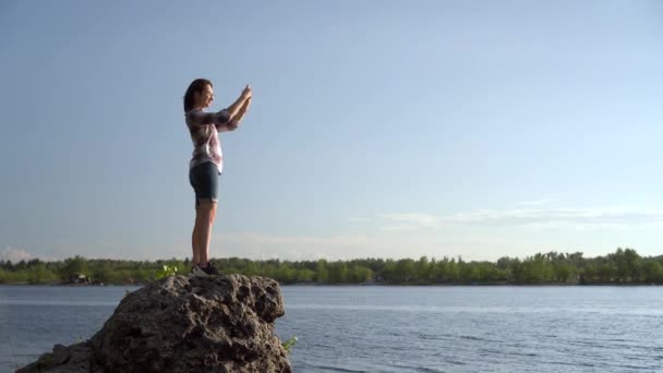 A young woman stands on a stone near the river and photographs the landscape. The girl takes pictures in nature. — Stock Video